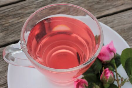 Tea rose herbal tea in a glass cup with pink buds and flowers on a rustic wooden table. Summer tea party outdoorsの写真素材