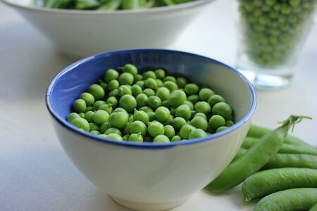 Fresh peeled green peas in a bowl and open pods on a white wooden table background with copy space. Top viewの写真素材