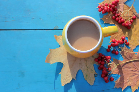 Flat lay composition with yellow cup of coffee, red berries, and autumn leaves on blue wooden background. Cozy autumn atmosphere. Top view, copy spaceの写真素材