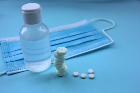 White pills, hand sanitizer bottle, and coronavirus medical disposable face mask on blue background...