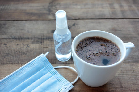 Cup of coffee, hand sanitizer bottle, and disposable medical face mask on wooden table from above....