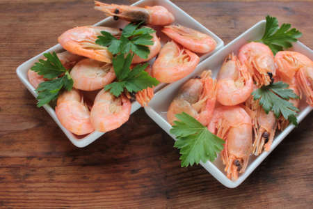 Cooked pink shrimps in the dish served with parsley. Shell-on North Atlantic prawns on the white plates on rustic wooden table. Top view, copy spaceの写真素材