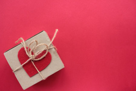 Craft gift box with tied with twine and decorated with handmade felt fabric heart on red background. Flat lay valentine`s day composition / Copy spaceの写真素材