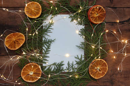 Christmas frame with Christmas tree branches, blank note white shit of paper, dried orange ornaments, and garland on vintage wooden background. Flat lay winter holiday composition.の写真素材