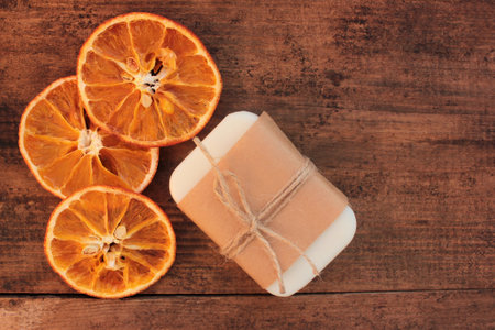 Citrus soap bar wrapped in craft paper and dried orange slices on wooden table. Handmade body care products. Top view, copy spaceの写真素材