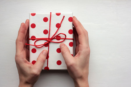 Person holding a gift box. Overhead view of female hands with present wrapped in red and white polka dot pattern paper on white background.の写真素材