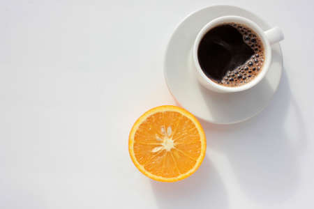 Cup of coffee and half of orange fruit on white background. Top view, copy space. Morning espresso on table. Healthy breakfast. Direct light, hard shadow.の写真素材