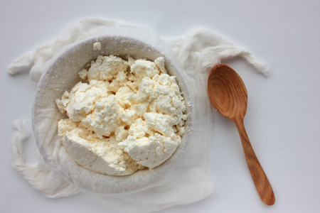 Close-up of freshly made white soft cottage cheese in bowl and wooden spoon on white table...