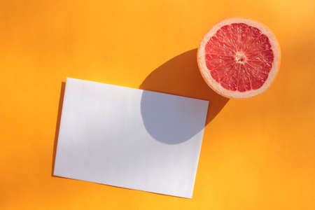 Summer still life with half of grapefruit and blank paper card on orange background. Invitation mockup on colored backdrop. Top view, copy pace. Hard light, shadowの写真素材