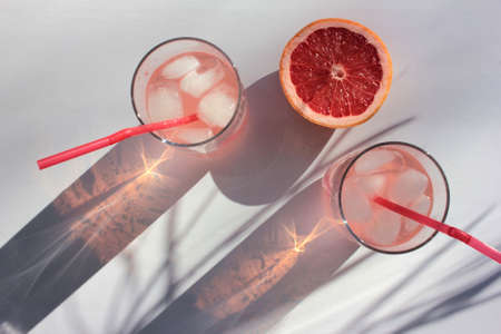Glass of pink drink with ice cubes and slice of grapefruit on white table background. Fruity refresh cocktails. Hard light. Palm leaves shadow. Top view. Summer party conceptの写真素材