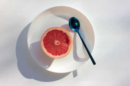 Half of ripe grapefruit on plate and spoon on white background. Flat lay food composition with bright citrus fruit on table. Hard light, shadow. Top view, copy spaceの写真素材