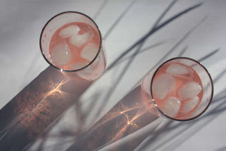 Glass of pink drink with ice cubes on white table background. Fruity refresh summer cocktails. Hard light. Palm leaves shadow. Top view.の写真素材