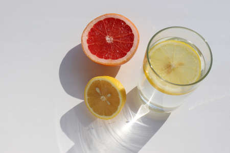 Glass of water with lemon slice and half of grapefruit on white background. Summer still life. Sunny day. Hard light, shadow. Top viewの写真素材