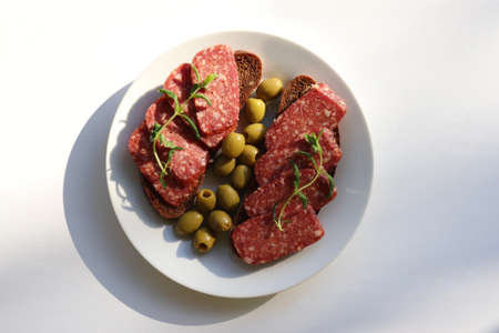 Salami pepperoni on rye bread sandwich with green pickled olives on plate on white table background. Flat lay food. Top view, copy spaceの写真素材