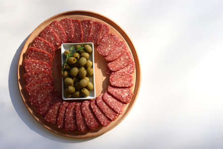 Slices of salami pepperoni and green pickled olives on round wooden cut board on white background. Top view, copy spaceの写真素材