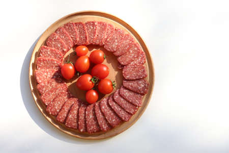 Slices of salami pepperoni and cherry tomatoes on round wooden cut board on white background. Top view, copy spaceの写真素材