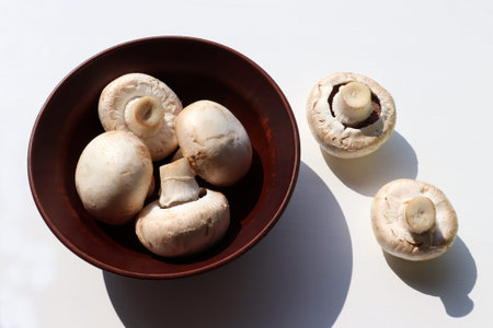 Raw champignon mushrooms in brown ceramic bowl on white table background. Top view, copy space. Hard light, shadowの写真素材