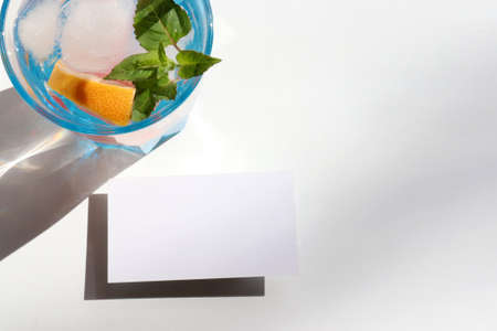 Summer cocktail with grapefruit and mint and blank paper card on white background. Refreshing drink with ice cubes and mock up invitation. Glass of lemonade with citrus fruit. Hard light, shadow.の写真素材