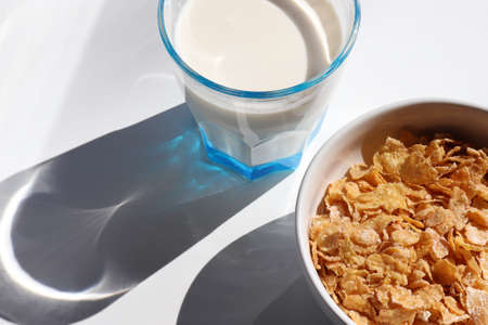 Bowl of cereal and glass of milk on white background. Healthy breakfast with cornflakes and milk. Top view, copy space. Hard light, shadowの写真素材