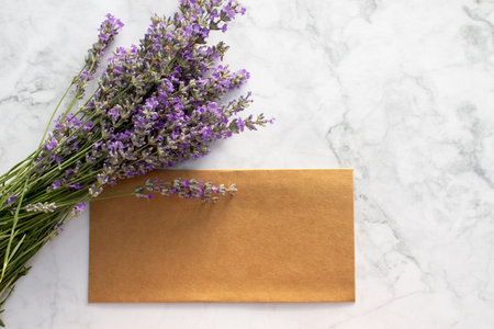 Summer stationery still life with lavender flowers and blank kraft paper envelope wedding invitation on white marble background. Mock up of invitation card. Top view, flat lay, copの写真素材