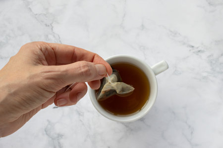 Person holding a tea bag. Teabag in woman hand. Cup of green herbal tea on white marble table...