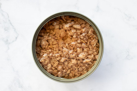 Open tin can with chopped tuna in oil on white marble kitchen table. Canned fish in a tin can. Top...