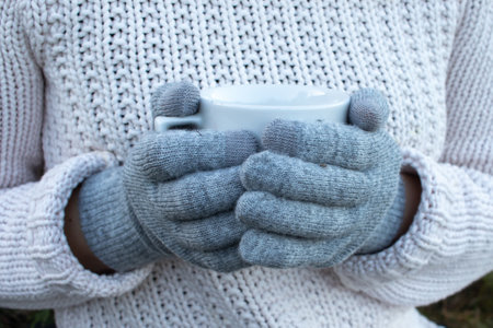 Hand holding a cup of tea, coffee, cocoa or hot chocolate. Woman in white knitted sweater and gray gloves holding cup of hot drink. Close upの写真素材