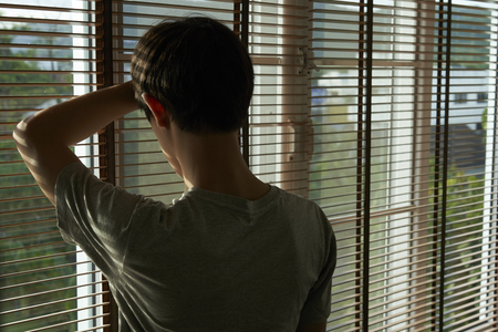 Depressed teenager standing and looking out the window. He has major depression and is unhappy.の写真素材