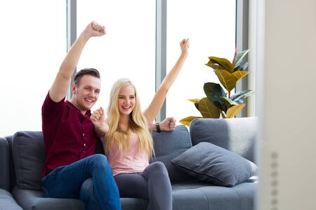 Young couple caucasian sitting in front of the television while cheering football happily, Handsome man and beautiful woman raised hands over the head because he was glad.の写真素材