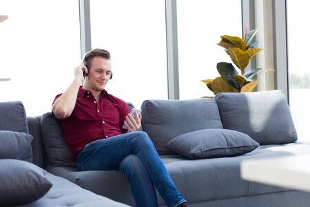 Adult handsome man wearing earphones listening to music from a smartphone while sitting on the sofa.の写真素材