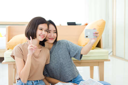 Two young Asian women enjoying taking selfie with mobile phones to update their status in online communities while resting at home.の写真素材