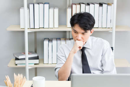 Asian male employee is stressed and thinking about work while sitting at the working desk inside the office.の写真素材