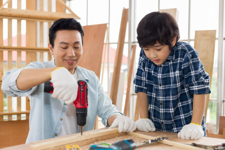 Asian father works woodwork while his son helps nearby.の写真素材