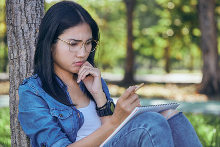 Asian female college student is sitting in the garden doing homework with a thoughtful expression.の写真素材