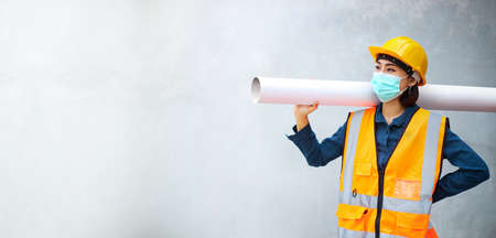 An engineer or a female worker wearing protective clothing and an anti-virus mask stood carrying a large pipe with a concrete wall in the background.の写真素材