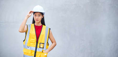 Portrait of an Asian female engineer or a construction worker wearing a yellow vest cloth stood and her hand touching a helmet, with a concrete background.の写真素材
