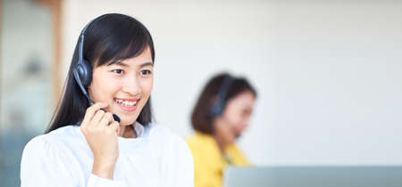 A beautiful young Asian woman wearing a headphone performs a call center function in a white office, blurred background of her co-workers.の写真素材