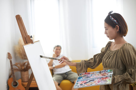 Asian woman who loves art or a female artist painter is holding a paint palette and paintbrushes while happily painting a portrait on white paper at studio.の写真素材