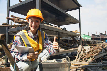 An Asian female factory worker wearing a hard hat and yellow vest sits holding a tablet, examining a used industrial battery with a pile of old material. Industrial waste management.の写真素材