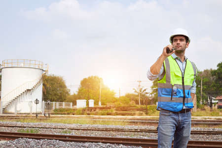 A handsome caucasian Railway engineer or Rail transport technician, wearing a reflective vest and helmet, stands and holding a walkie-talkie on an outdoor train track.の写真素材