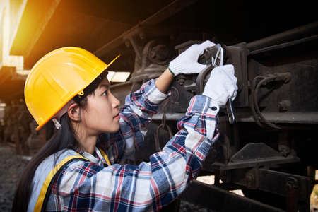 An Asian female maintenance worker wearing a helmet and yellow vest is using a wrench to repair the undercarriage of a freight train, adding orange lights.の写真素材