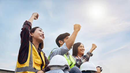Three Successful engineers or Industrial workers wearing safety vests were raising his fist to show joy and confidence in the outdoors with a beautiful sky and sun as a backdrop.の写真素材