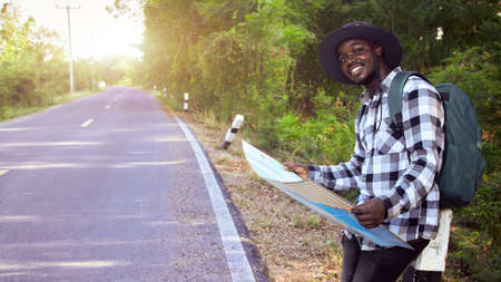 An African man in a long-sleeved shirt and a hat, holding a map and carrying a backpack, is standing on a rural road on a summer vacation. Ideas of travel and leisure time during the holidays.の写真素材