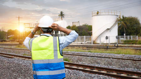 The back of a caucasian Railway engineer or Rail transport technician, wearing a reflective vest and helmet, standing outdoors in the morning when the sun is rising.の写真素材