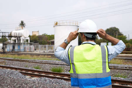 The back of a caucasian Railway engineer or Rail transport technician, wearing a reflective vest and helmet, standing outdoors in the morning.の写真素材