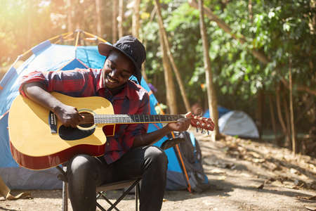 Afro man wearing casual clothing sits comfortably in a chair in front of the tent enjoying playing acoustic guitar. Young male tourists spend their vacations doing hiking and camping activities.の写真素材