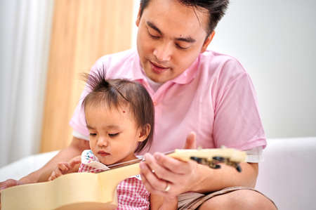 Two year old daughter playing guitar with father on the sofa. A single dad is taking care of a little daughter. Skills training and child development, family relationships.の写真素材