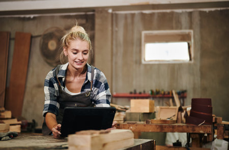 Caucasian female carpenter wearing an apron is calculating and drawing woodwork on digital tablet while working at her workplace.の写真素材