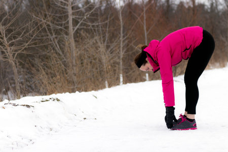 Sporty fit woman stretches her hamstrings before running on trail on cold winter day. Wellness...