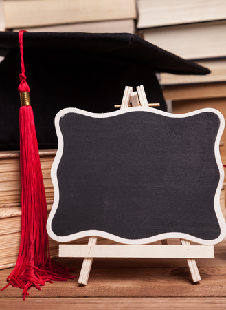 Little blackboard next to a black mortarboard with red tassel and some booksの写真素材